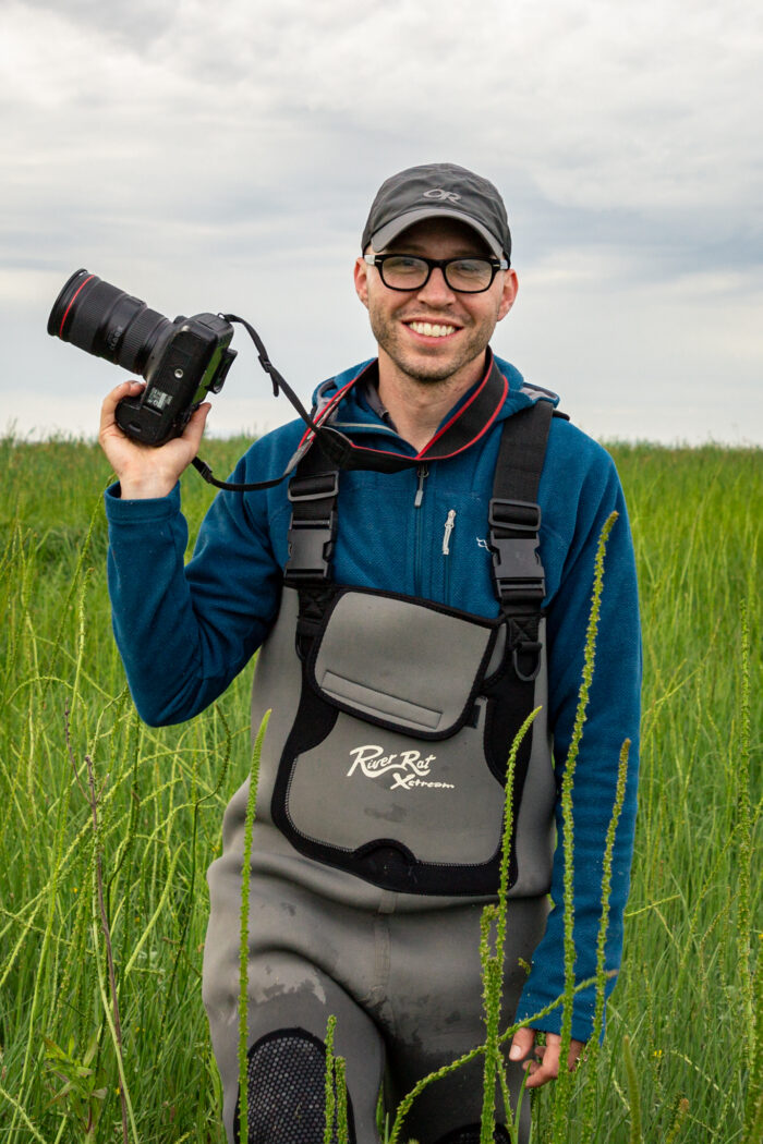 A portrait photo of a white man standing in a grassy field. He is holding a camera and wearing a chest wader. He is smiling.