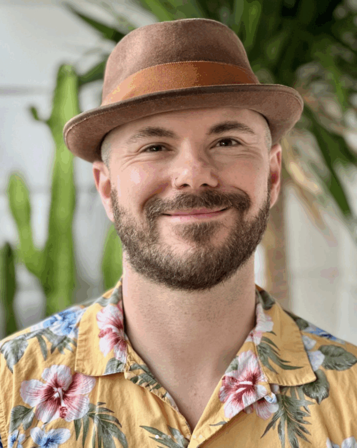 A person with a beard, hat and nose piercing smiles toward the camera for a portrait photo.