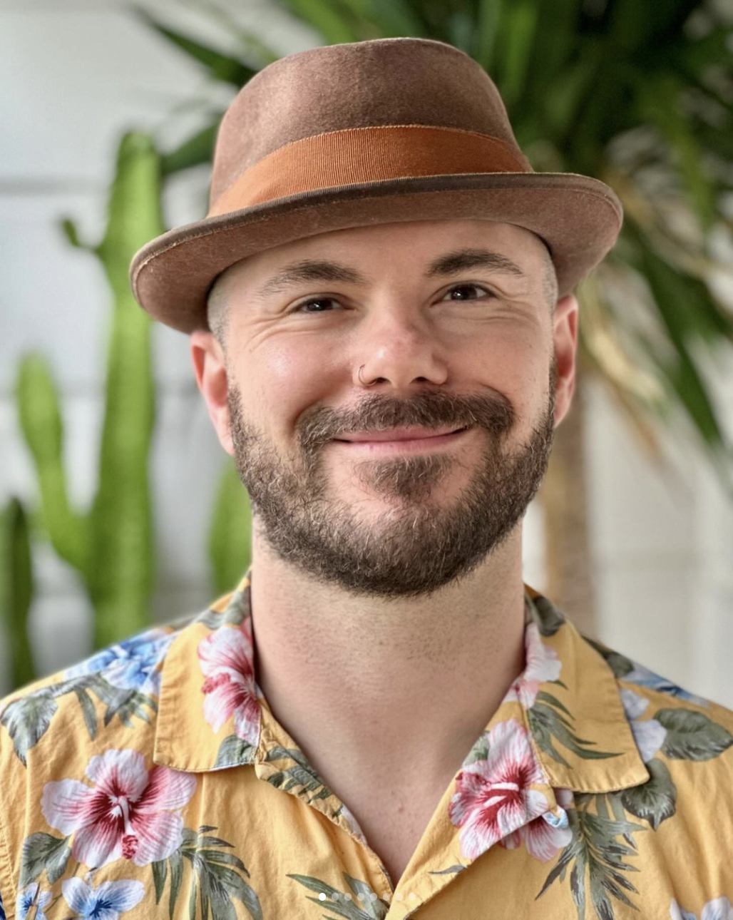 A person with a beard, hat and nose piercing smiles toward the camera for a portrait photo.