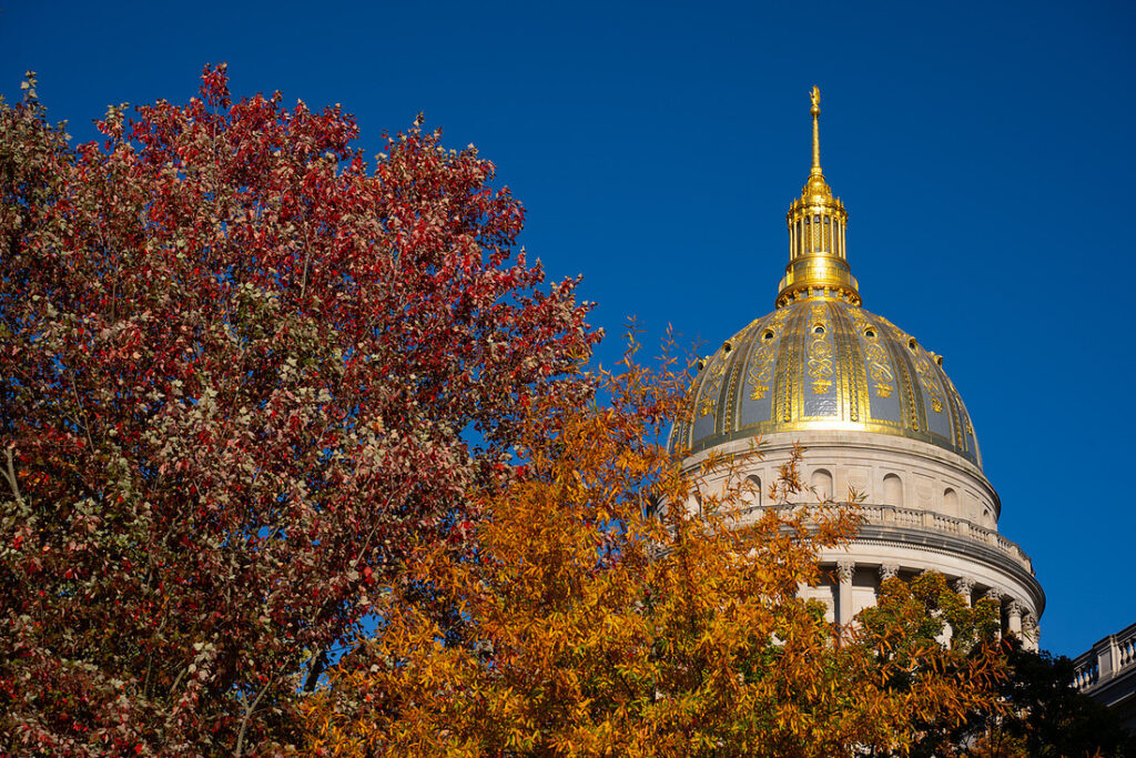 The gilded top of the West Virginia State Capitol is visible in the sky beside autumnal leaves changing colors on the treetops.