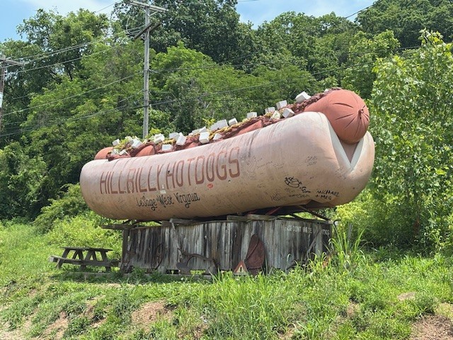 A giant hotdog sign advertising HillBilly Hotdogs in LeSage sits on top of a rough looking wooden platform next to a picnic table.