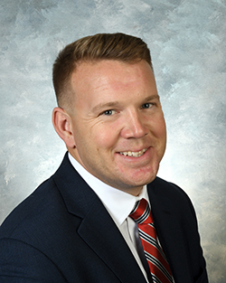 A professional headshot of a middle age white man smiling toward the camera. He has short brown hair and wears a navy blazer, white button up shirt and red striped tie.