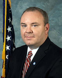 A professional headshot of a middle age white man. He has brown hair and wears a black blazer, white button up shirt, and a red striped tie. His hair is trimmed very short. Behind him is the American flag.