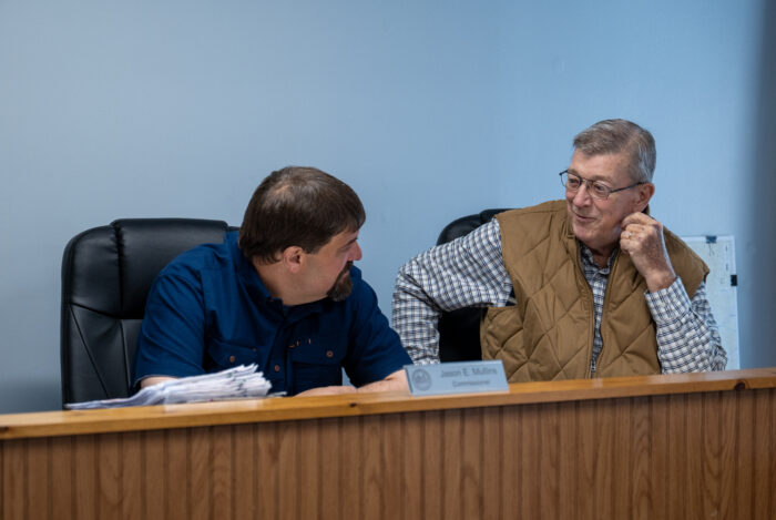 Two white men sit behind a wooden wall talking. 