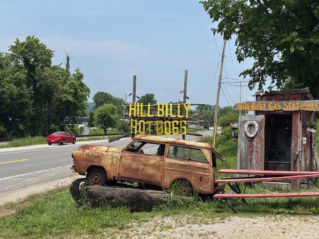 A junked car advertising Hillbilly Hotdogs in LeSage sits in front of an outhouse and overlooks the road.