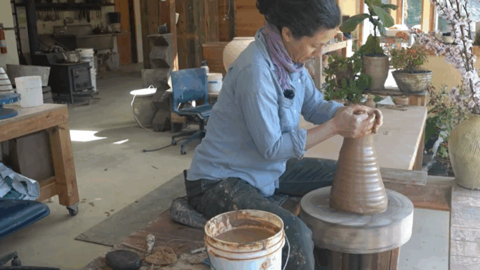 A woman molding a piece of clay into a vase. She is working in a studio with other finished pieces of pottery.