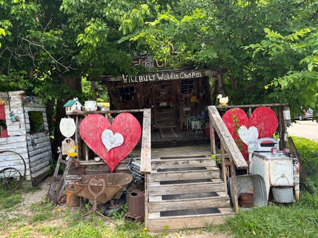 A rustic looking wedding chapel sits under a tree. Big wooden hearts decorate the porch.