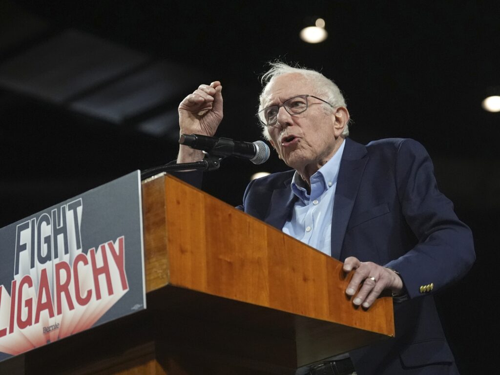 A man stands at a wooden lectern with a sign that reads "Fight Oligarchy" pasted to the front. The man, with thinning white hair and wire frame glasses, wears a blue blazer over a light blue Oxford button up shirt. He gesticulates with his right hand while speaking into a wireless microphone.
