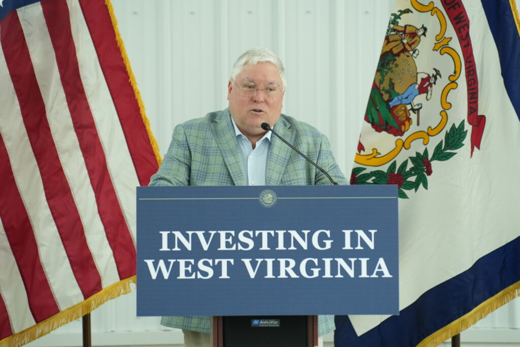 A man stands at a lectern with a sign that reads "Investing in West Virginia" between a U.S. flag and a West Virginia flag. He wears a grey checkered jacket over a white button up