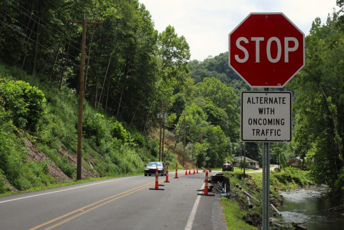 A stop sign with a black and white sign below that reads "Alternate with oncoming traffic" stands to the right of a road covered in thin, orange plastic bollards. The bollards block off a section of road that has collapsed over a river embankment. To the left of the bollards, a car drives towards camera on the open lane of traffic.