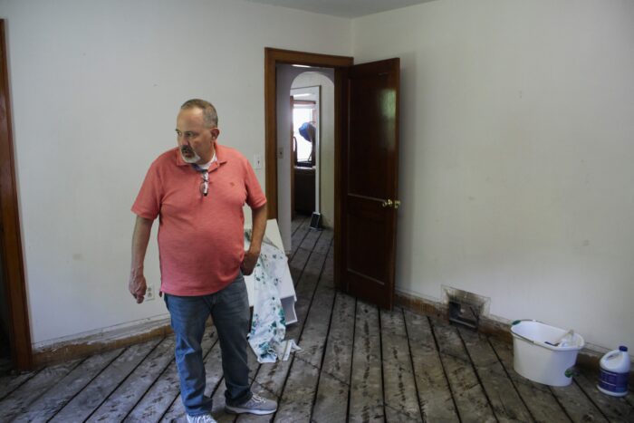 A man in a salmon polo and blue jeans looks off camera left. He stands in an empty room with exposed subfloor slats. Behind him, more empty rooms and stripped floors are visible further into the building.