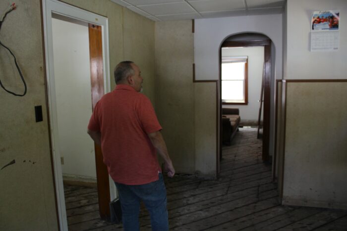 A man in a salmon polo and blue jeans looks at a calendar on a wall. He stands in an empty room with exposed subfloor slats.