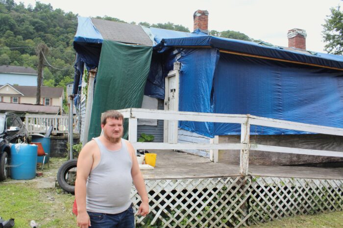 A man in a white tank top and jeans stands in front of a house covered in green and blue tarps.