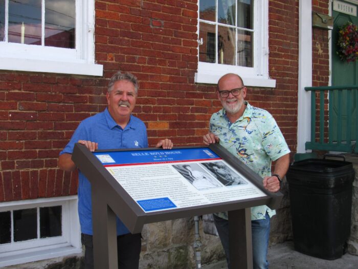 Two men stand beside a historical information sign outside a red brick building. They are smiling and looking toward the camera.