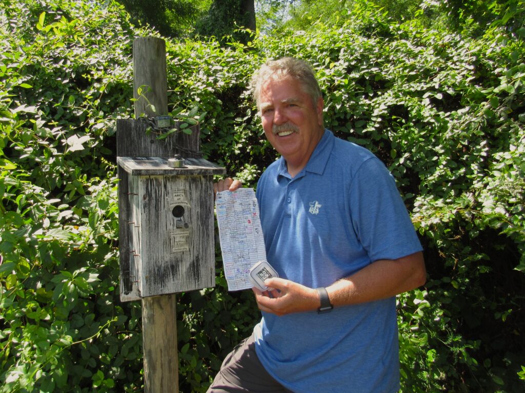 A man in a blue polo shirt stands beside a bird house and holds a sheet of paper with numerous colorful signatures on it. He smiles and looks toward the camera.