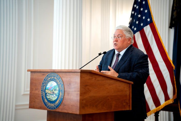 Man in dark suit stands behind wooden podium