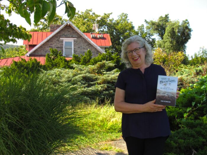 A woman holds a book and smiles at the camera. She stands before an old stone building and a lush yard with lots of shrubs.