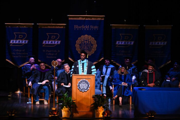 A man in university regalia including a black tam with a gold tassel and a black and turquoise robe stands at a wooden lectern with the Bluefield State University seal on its front. He stands in front of a dark blue upholstered seat and a dais of men and women in similar academic regalia on a stage. Above all of them are hung five large pendants with Bluefield State branding, an outlined B with a 'state' banner in front of it, in dark blue and gold.