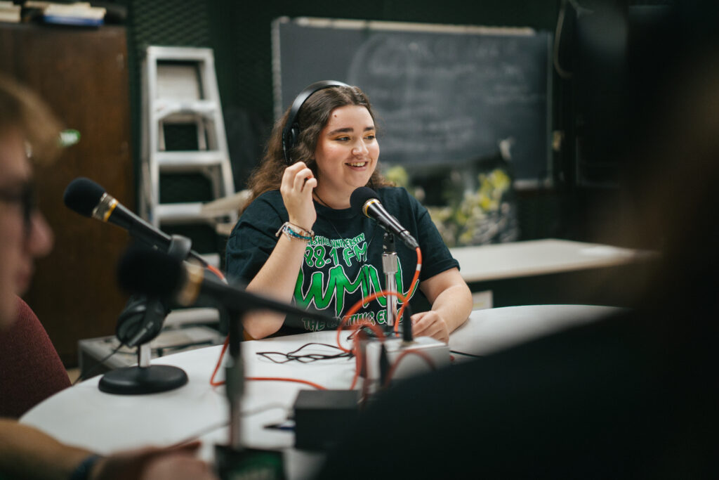 A young woman wears headphones and a black shirt with green text that reads "88.1 FM WMUL" speaks into a microphone in a table stand with a red XLR cable feeding away from it. Behind the woman can be seen a blackboard.