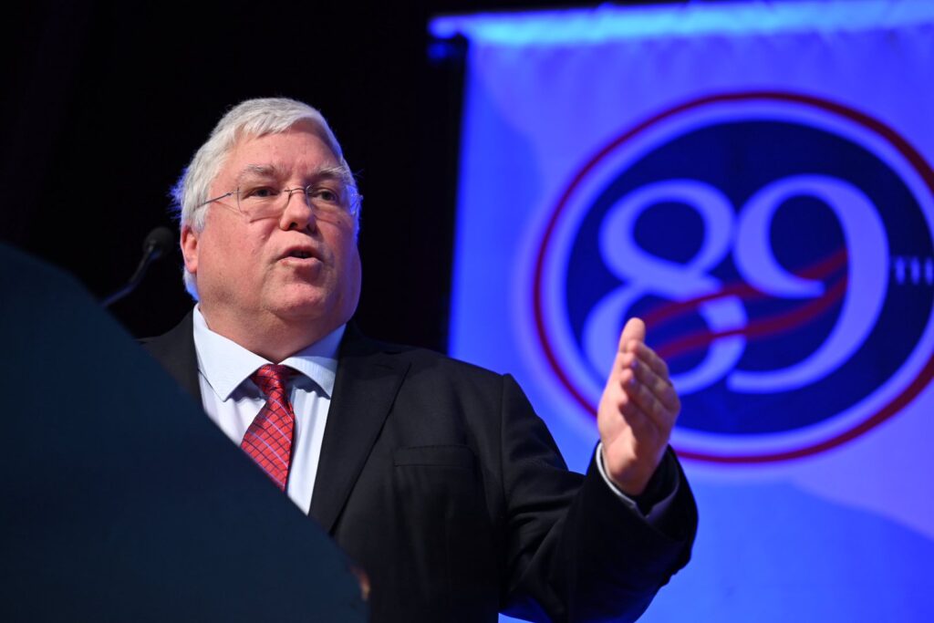 A man with white hair wearing wireframe glasses and a black suit over a white collared shirt with a red striped tie gestures with his left hand. He stands behind a dark blue lectern and in front of a large white banner with an 89 in a blue circle.