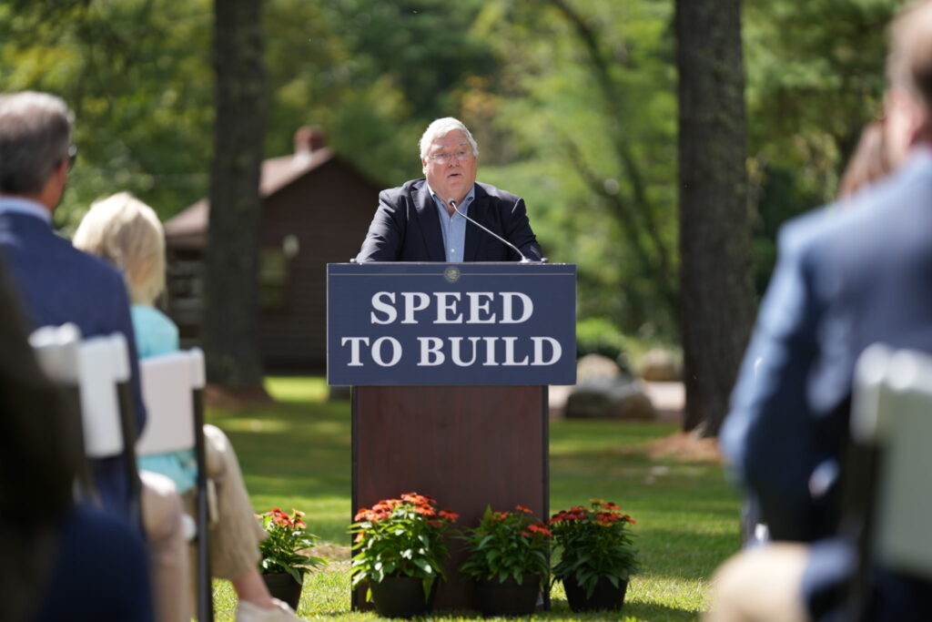 man in blue suit standing behind a podium that says speed to build