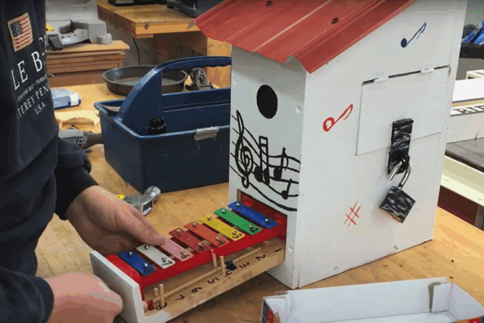 A rainbow-colored xylophone protrudes from the bottom of a birdhouse placed on a wooden work bench.