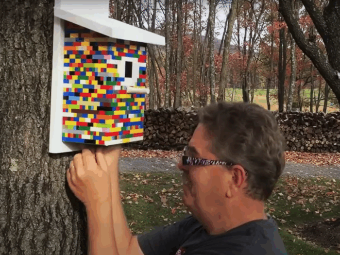 A man fiddles with a birdhouse made of colorful legos hanging on a tree.