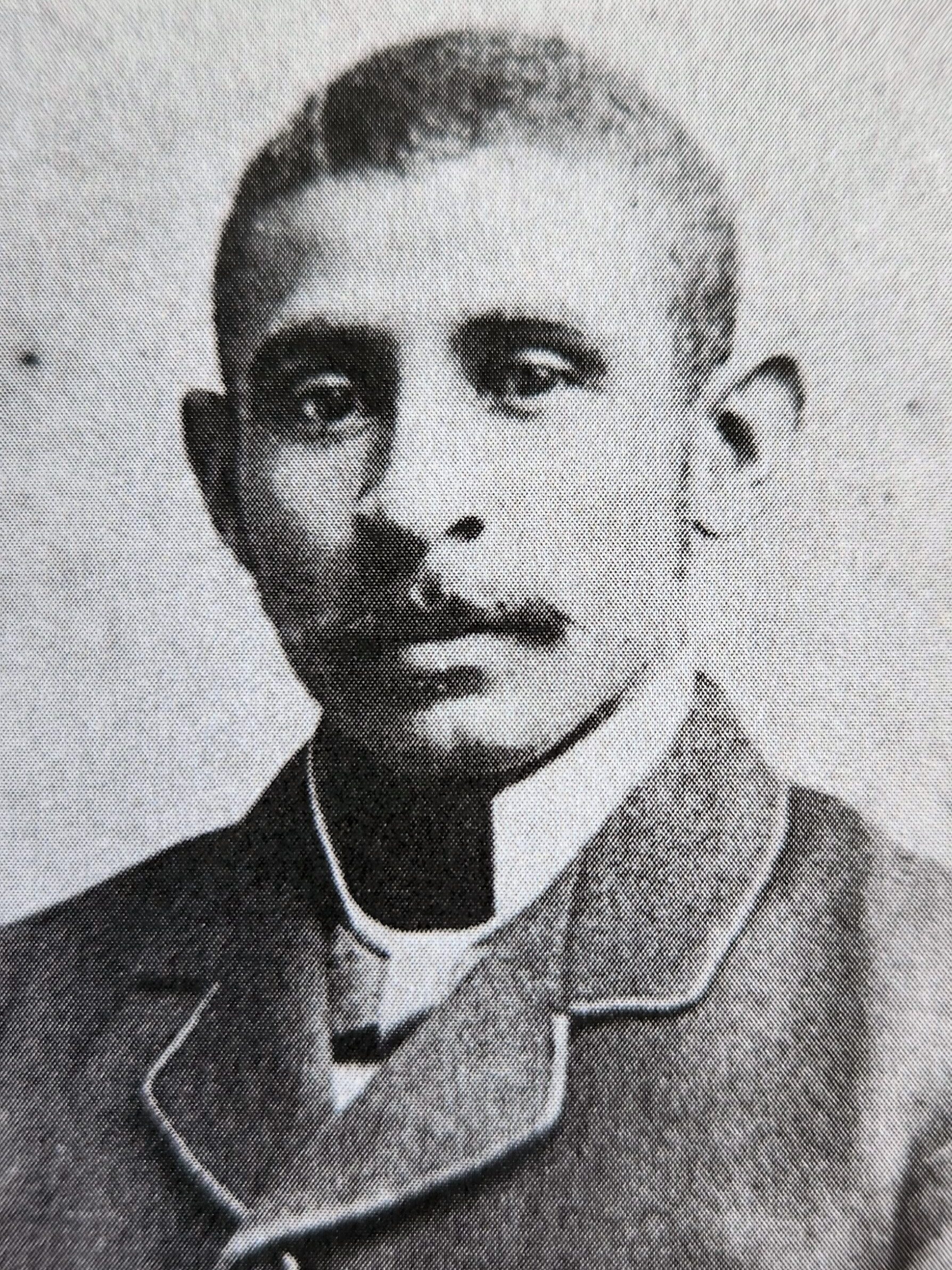 A Black man in a formal attire sits for a headshot photo and looks at the camera. The photo is black-and-white and visibly old.
