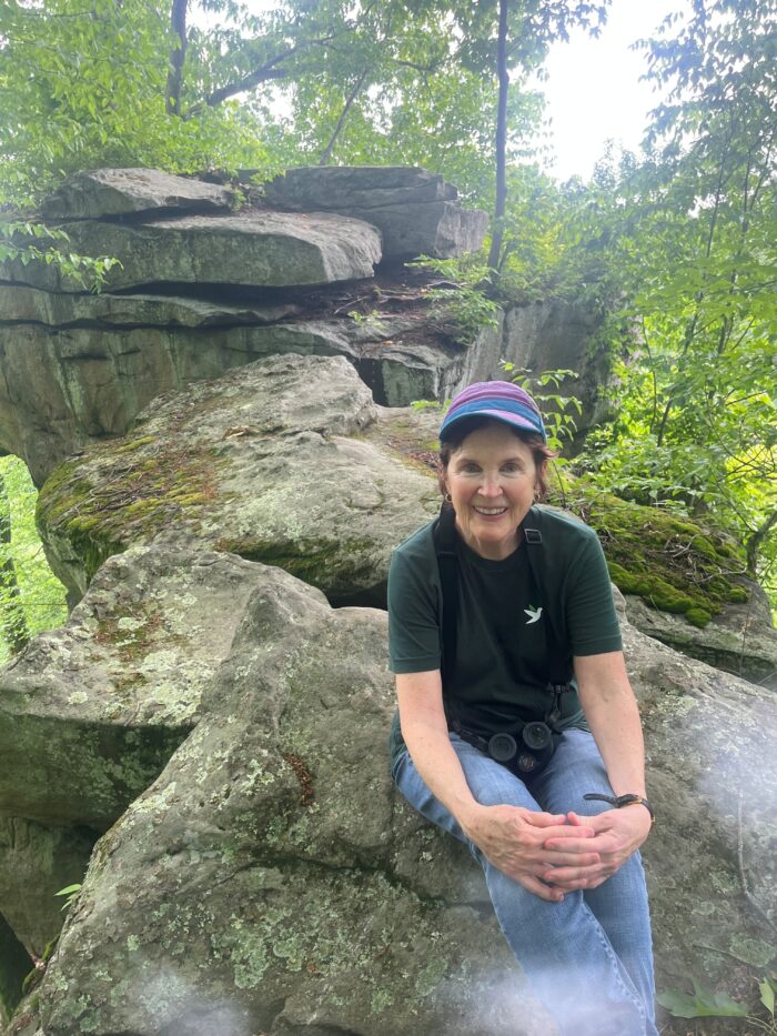 An older white woman sitting on a rock surrounded by trees.