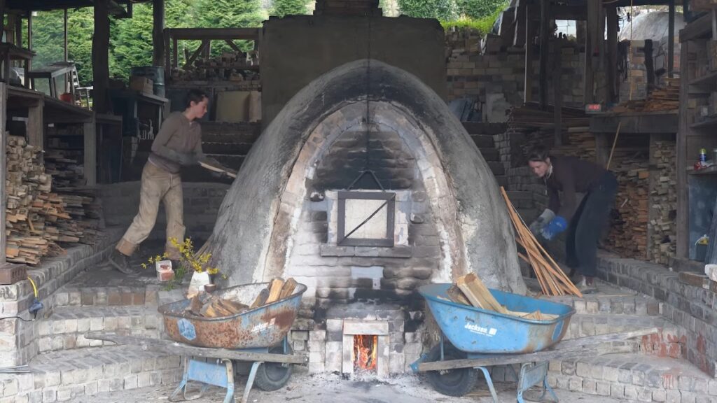 In the center, there is a wood fired kiln. There are two blue wheelbarrows in front of the kiln. There are also two people scooping something into the kiln with a shovel.