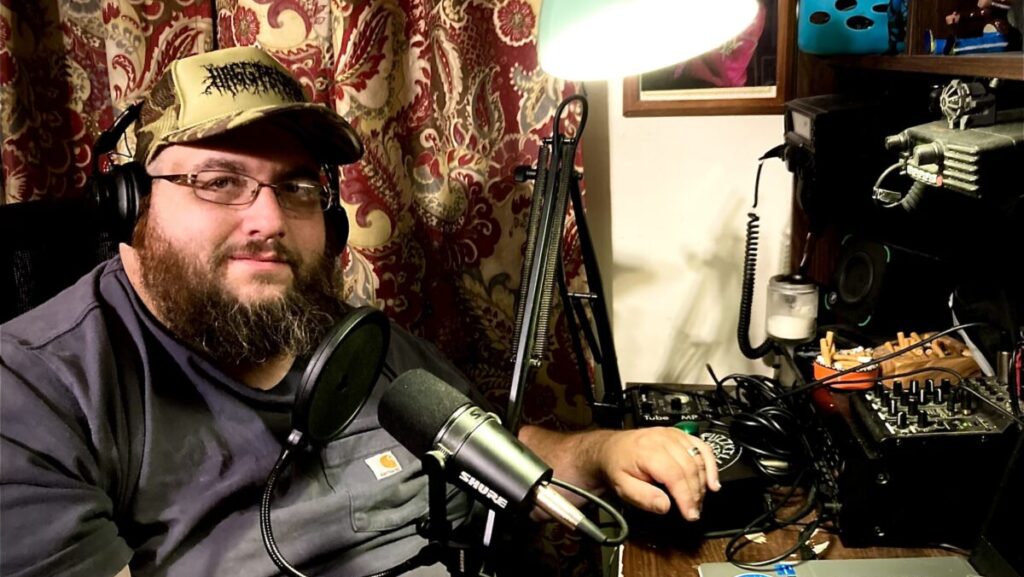 A white man sitting at a desk. There is a microphone and a lamp in front of him.