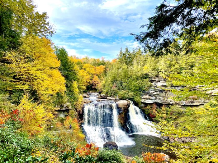 A photo of a waterfall with trees on every side. The leaves are turning colors for fall. It is during the day with some clouds in the sky.