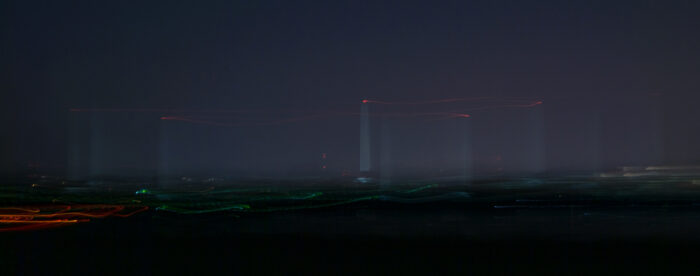 A photograph at nighttime. Tall towers are seen in the distance. Red lights. Blurred lights.