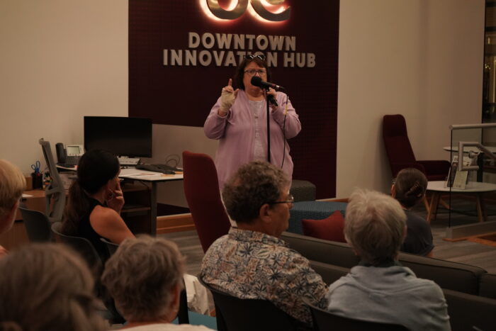 A white woman wearing a light purple sweater speaks into a microphone. She has a wrap on her hand, sunglasses on top of her head, and wears glasses. Audience members look on.