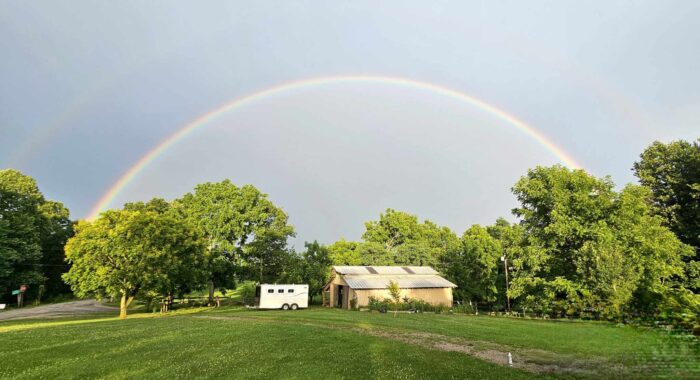 A faraway photo of a rainbow over a house. Green trees and green grass. Blue sky.