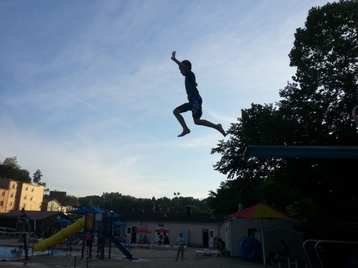 A silhouette of a young man jumping off a high dive against a blue sky. 