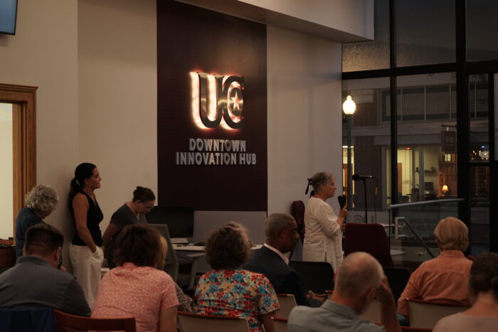 About a dozen people are seen in an audience at the UC Downtown Innovation Hub. A woman speaks into a microphone as three women wait in a line behind her for their turn to speak.