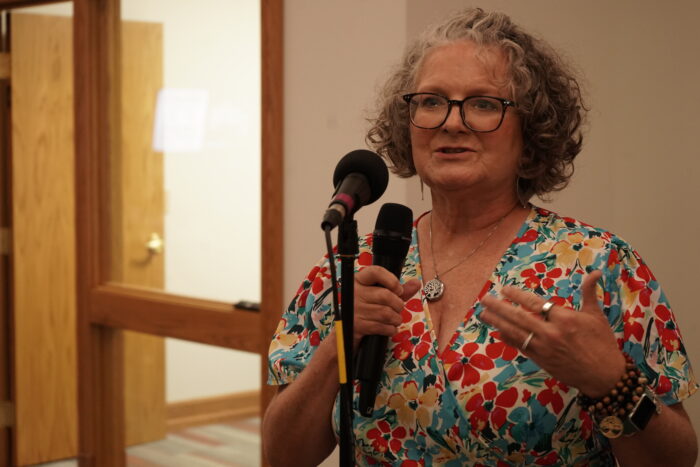 An older white woman with gray hair, wearing glasses, a tree of life necklace and a colorful dress, speaks into a microphone.
