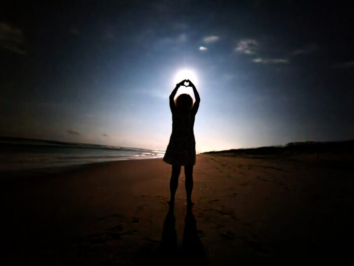 The silhouette of a woman against a moon in the sky. She holds her hands up in a heart shape against the moon. 