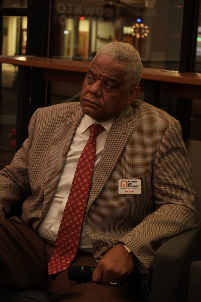 A Black man with short, gray hair looks off camera at a speaker. He wears a tan blazer, white button up shirt, and red tie. He holds a microphone.