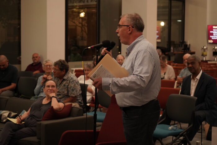 An older white man with gray-white hair speaks into a microphone at an event. In the background are audience members listening.
