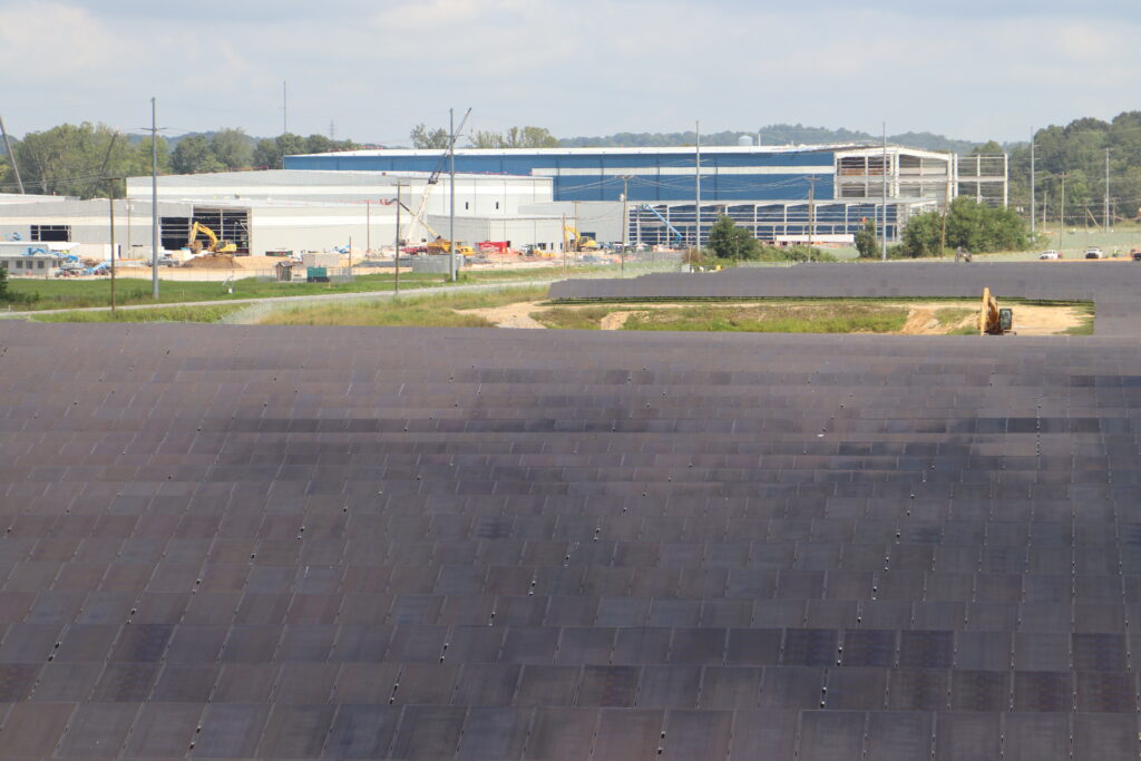 A field of solar panels reflect the sky on a partly cloudy day. A blue industrial building rises in the distance.