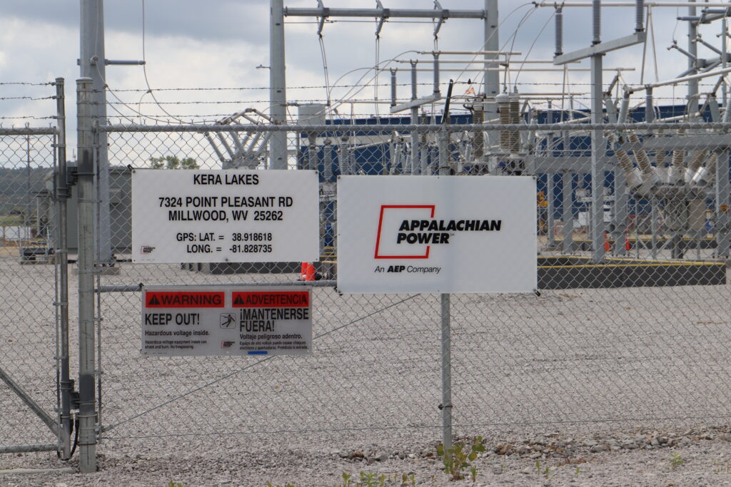 An electrical substation sits behind a fence with signs attached to it on a partly overcast day.