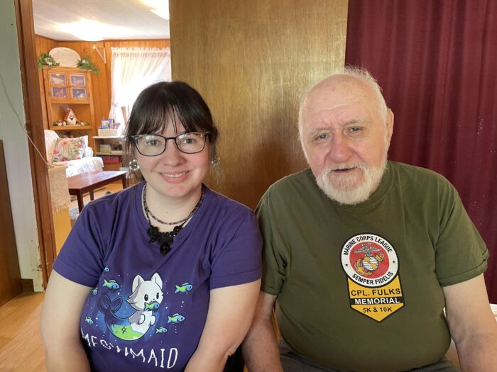 Two people, a white woman and an older white man. Daughter and father. The woman wears a purple t-shirt featuring a mermaid cat cartoon character. The man wears a green shirt with a Marine Corps League logo. They are sitting in their home.