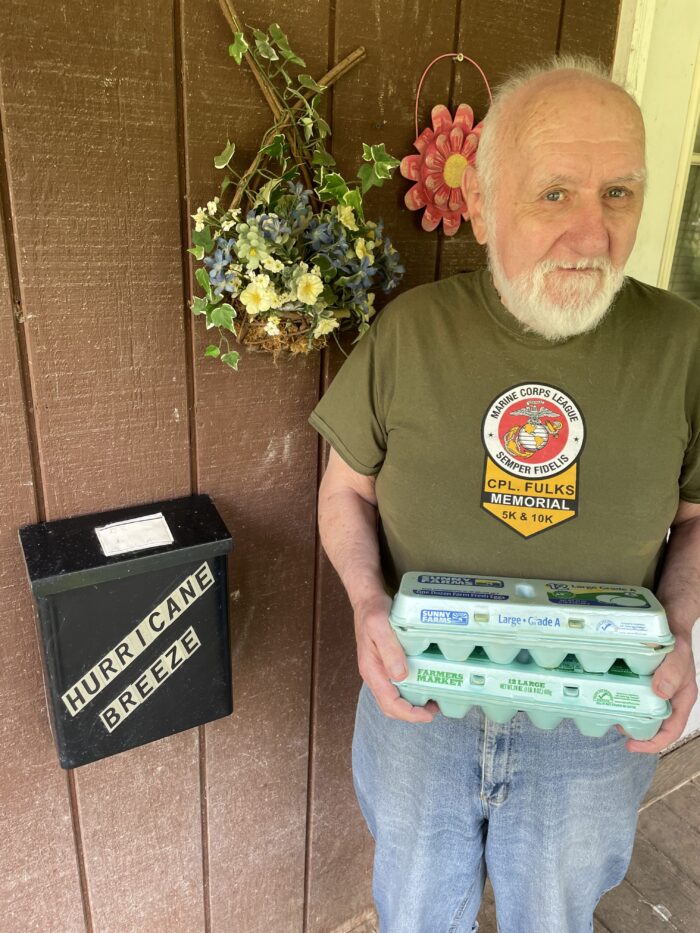 An older white man stands in front of a door holding two dozen eggs in cartons.