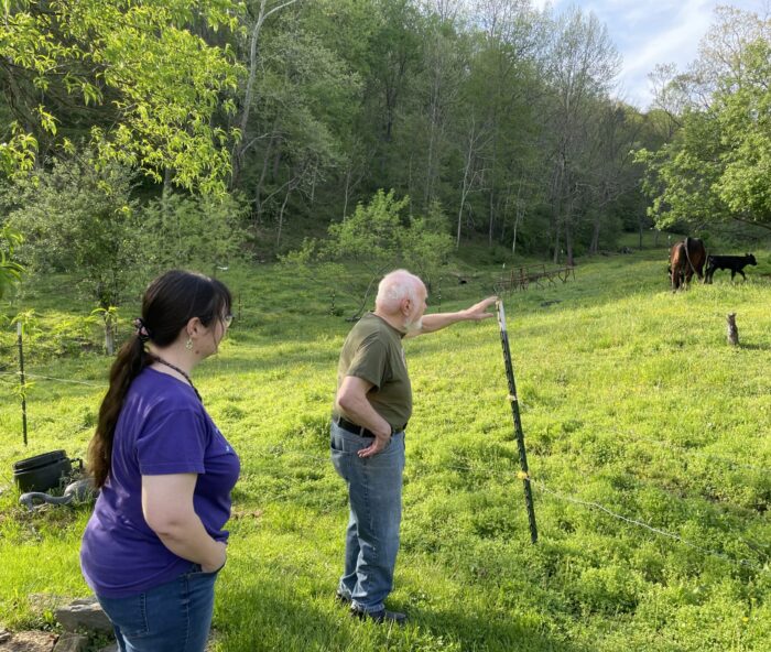 Two people, a white woman and an older white man, daughter and father, stand outside looking off at farm animals.