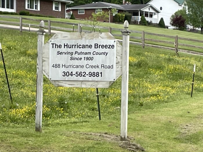A sign is seen on a grassy hillside. The sign reads, "The Hurricane Breeze, Serving Putnam County Since 1900. 488 Hurrican Creek Road. 304-562-9881."