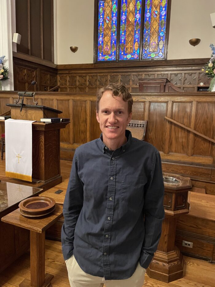 A white man stands in a church smiling for the camera. He wears a dark button up shirt and slacks.