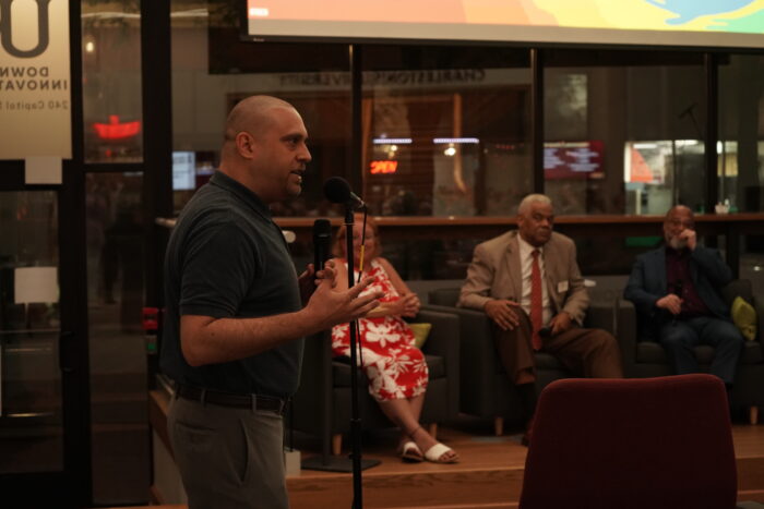A white, bald man wearing a dark polo shirt and slacks speaks into a microphone.