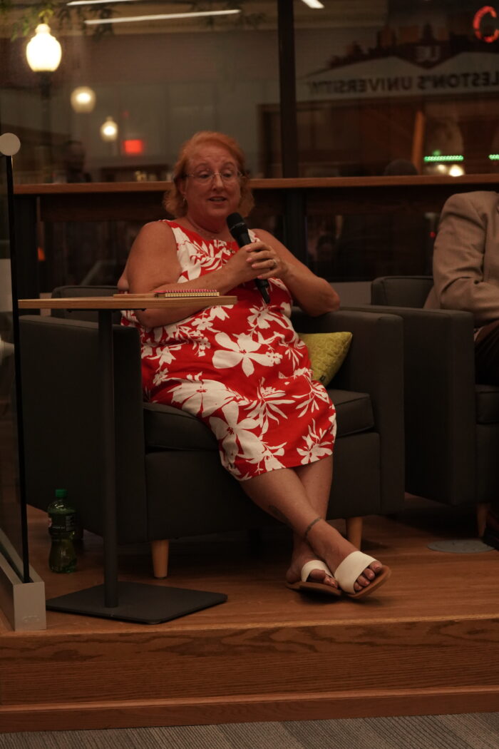 An older white woman with red hair wearing a red and white Hawaiian-style dress sits in a chair holding a microphone. She wears glasses.